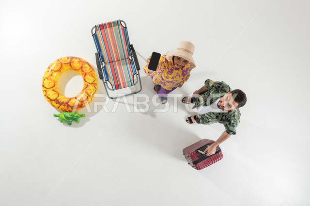 استخدام الأجهزة التقنية للترفيه في الإجازة , قضاء العطلة الصيفية على الشاطئ , بورتريه علوي عامودي لزوجان عربيان خليجيان سعوديان يرتديان ملابس الشاطئ الصيفية , عرض شاشة سوداء فارغة عبر الجوال , خلفية بيضاء