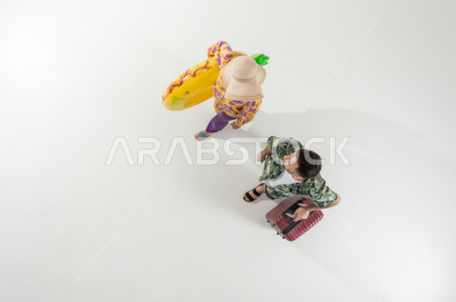 الاستعداد للفعاليات الصيفية الممتعة , بورتريه علوي عامودي لزوجان عربيان خليجيان سعوديان يرتديان ملابس الشاطئ الصيفية يحملان حقيبة سفر و عوامة السباحة , السفر و الرحلات في السعودية , خلفية بيضاء