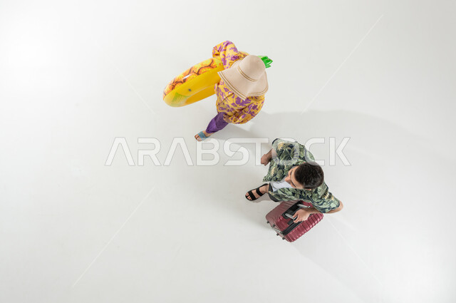 Leisure activities in Saudi Arabia, preparing for a beach trip on vacation, a vertical overhead portrait of a Saudi Gulf Arab couple wearing summer beach clothes, carrying a travel bag and a swimming buoy, white background
