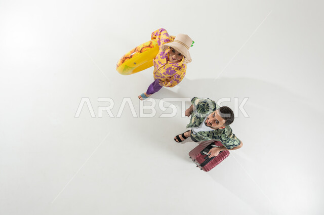 فعاليات و رحلات في الإجازة , بورتريه علوي عامودي لزوجان عربيان خليجيان سعوديان يرتديان ملابس الشاطئ الصيفية يحملان حقيبة سفر و عوامة السباحة , الذهاب إلى الشاطئ في العطلة الصيفية, خلفية بيضاء