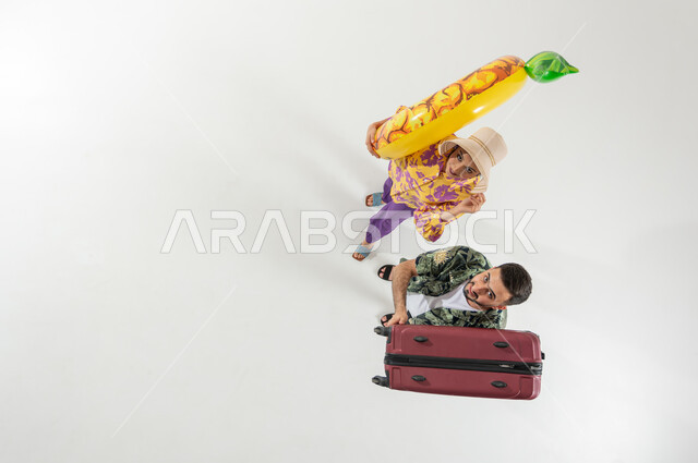 الاستعداد لقضاء الإجازة على الشاطئ , بورتريه علوي عامودي لزوجان عربيان خليجيان سعوديان يرتديان ملابس الشاطئ الصيفية يحملان حقيبة سفر و عوامة السباحة , النظر إلى الكاميرا بإيماءات الفرح و السرور ,خلفية بيضاء