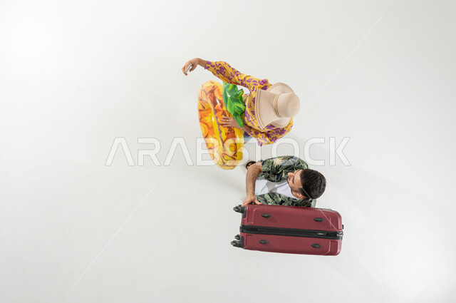 التحضير لرحلات الشاطئ في الإجازة , بورتريه علوي عامودي لزوجان عربيان خليجيان سعوديان يرتديان ملابس الشاطئ الصيفية يحملان حقيبة السفر و عوامة السباحة , الاستمتاع بالعطلة الصيفية , خلفية بيضاء