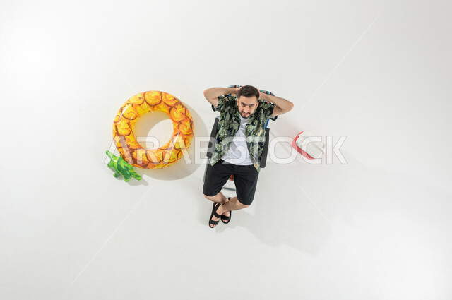 الاسترخاء و الاختلاء على الشاطئ , الاستمتاع بالعطلة الصيفية , بورتريه علوي عامودي لرجل عربي خليجي سعودي يرتدي ملابس الشاطئ الصيفية يجلس على الكرسي , النظر إلى الكاميرا بإيماءات الفرح و السرور , خلفية بيضاء