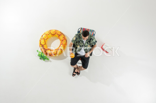 الاستجمام و الاختلاء في العطلة الصيفية , قضاء الإجازة على الشاطئ , بورتريه علوي عامودي لرجل عربي خليجي سعودي يرتدي ملابس الشاطئ الصيفية يجلس على الكرسي يحمل كأس العصير بيده , خلفية بيضاء