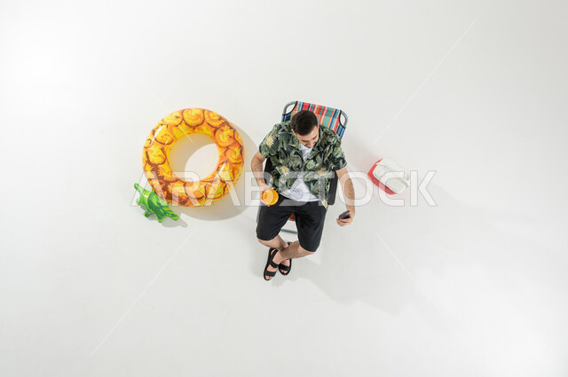 مشاركة الأصدقاء اللحظات الممتعة عن طريق مكالمة الفيديو الهاتفية , بورتريه علوي عامودي لرجل عربي خليجي سعودي يرتدي ملابس الشاطئ الصيفية يجلس على الكرسي يحمل الهاتف المحمول و كأس العصير بيديه  , خلفية بيضاء