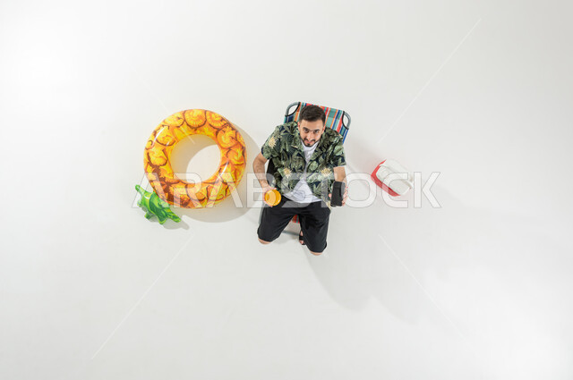 استخدام التكنولوجيا للتسلية و الترفيه , قضاء إجازة الصيف على الشاطئ , بورتريه علوي عامودي لرجل عربي خليجي سعودي يرتدي ملابس الشاطئ الصيفية يجلس على الكرسي و يحمل الهاتف المحمول و كأس العصير بيديه , عرض شاشة سوداء فارغة عبر الجوال , خلفية بيضاء