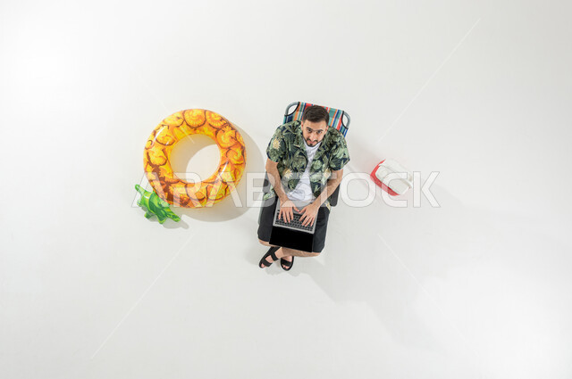 استخدام الأجهزة الحديثة للتسلية في العطلة الصيفية , بورتريه علوي عامودي لرجل عربي خليجي سعودي يرتدي ملابس الشاطئ الصيفية يجلس على الكرسي يقوم بالكتابة على لوحة مفاتيح الحاسب المحمول  , عرض شاشة سوداء فارغة عبر الحاسوب , خلفية بيضاء