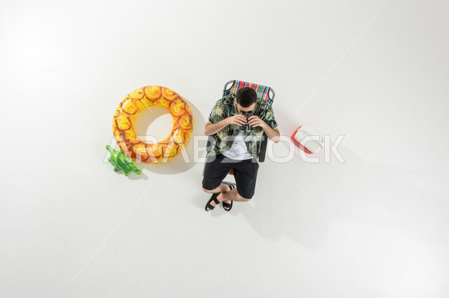 عطلة صيفية ممتعة , الاستمتاع بالإجازة على الشاطئ , بورتريه علوي عامودي لرجل عربي خليجي سعودي يرتدي ملابس الشاطئ الصيفية يجلس على الكرسي يقوم بالنظر من خلال المنظار ثنائي العينين , خلفية بيضاء