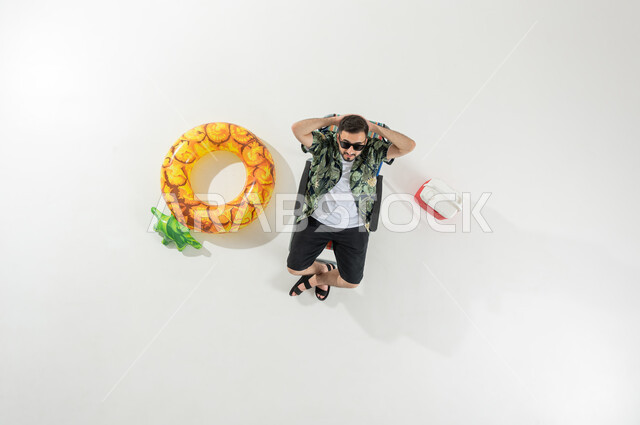 رحلة الشاطئ الممتعة , بورتريه علوي عامودي لرجل عربي خليجي سعودي يرتدي ملابس الشاطئ الصيفية مع نظارات شمسية يجلس على كرسي الاسترخاء , النظر إلى الكاميرا بإيماءات الراحة و السعادة , خلفية بيضاء