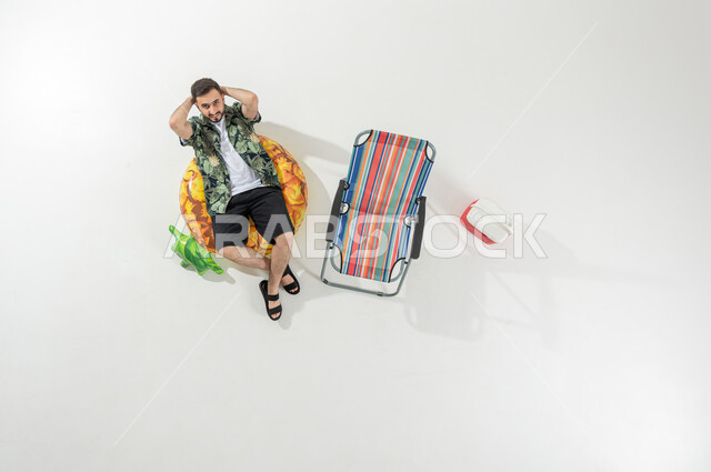 قضاء إجازة الصيف على الشاطئ , بورتريه علوي عامودي لرجل عربي خليجي سعودي يرتدي ملابس الشاطئ الصيفية يستلقي على عوامة السباحة , إيماءات الاسترخاء و السعادة , خلفية بيضاء