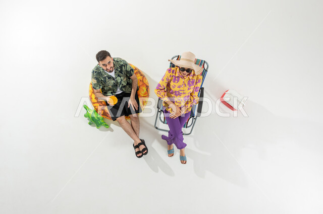 رحلة الشاطئ الممتعة , الاستمتاع بالمشروبات الباردة و المنعشة , بورتريه علوي عامودي لزوجان عربيان خليجيان سعوديان يرتديان ملابس الشاطئ الصيفية , النظر إلى الكاميرا بإيماءات الفرح و السرور , خلفية بيضاء