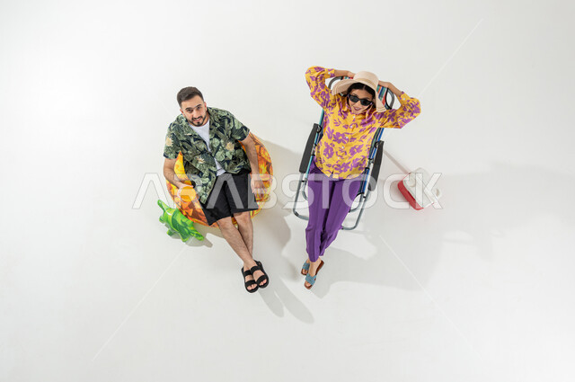 Enjoying cold and refreshing drinks, looking at the camera with relaxed and happy gestures, vertical overhead portrait of a Saudi Gulf Arab couple wearing summer beach clothes, spending vacation on the beach, white background