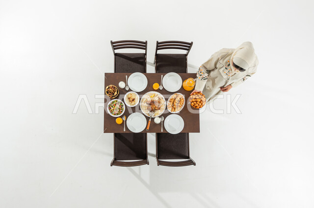 تحضير سفرة الإفطار بالمأكولات الشرقية , بورتريه علوي عامودي لامرأة عربية خليجية سعودية ترتدي الثوب التقليدي و الحجاب تقوم بتحضير مائدة الطعام , أجواء رمضانية في السعودية ,خلفية بيضاء