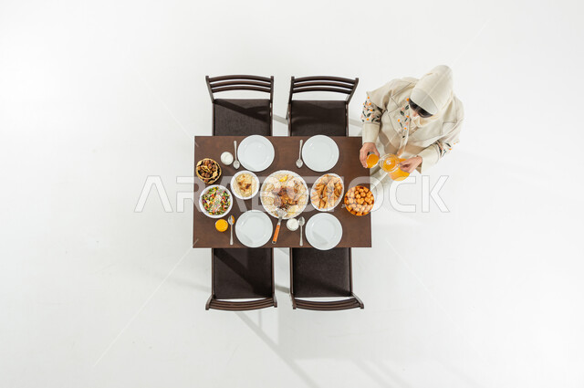 تقديم المشروبات الباردة على مائدة الإفطار , بورتريه علوي عامودي لامرأة عربية خليجية سعودية ترتدي الثوب التقليدي و الحجاب تقوم بتحضير مائدة الطعام , أجواء رمضانية في السعودية ,خلفية بيضاء