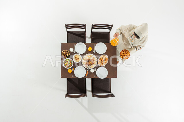 تقديم المأكولات الشرقية على مائدة الإفطار , بورتريه علوي عامودي لامرأة عربية خليجية سعودية ترتدي الثوب التقليدي و الحجاب تقوم بتحضير سفرة الطعام , أجواء شهر رمضان المبارك في السعودية ,خلفية بيضاء