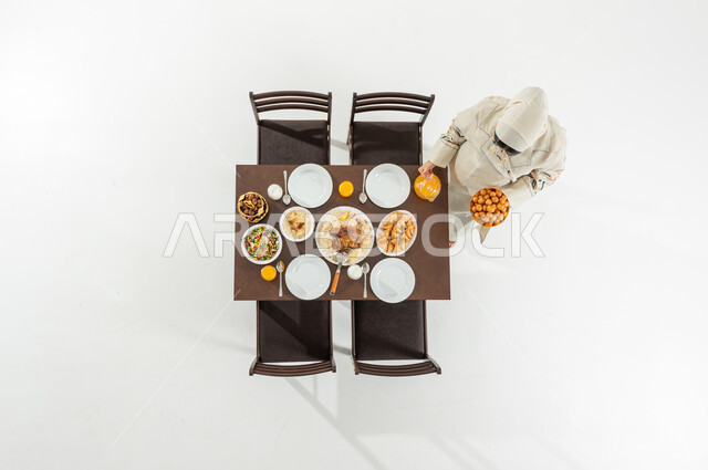 تحضير مائدة الإفطار , أجواء شهر رمضان المبارك , بورتريه علوي عامودي لامرأة عربية خليجية سعودية ترتدي الثوب التقليدي و الحجاب تقوم بتحضير سفرة الطعام , مأكولات شرقية شهية , أجواء رمضانية في السعودية ,خلفية بيضاء