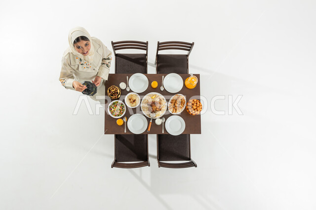 تجهيزات سفرة الإفطار , الفرح بقدوم شهر رمضان المبارك , بورتريه علوي عامودي لامرأة عربية خليجية سعودية ترتدي الثوب التقليدي و الحجاب تقوم بحمل فانوس رمضان , النظر إلى الكاميرا بإيماءات السعادة و السرور , خلفية بيضاء