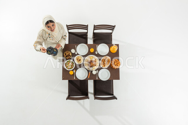 الفرح بقدوم شهر رمضان المبارك , تجهيزات سفرة الإفطار , بورتريه علوي عامودي لامرأة عربية خليجية سعودية ترتدي الثوب التقليدي و الحجاب تقوم بحمل فانوس رمضان , النظر إلى الكاميرا بإيماءات السعادة و السرور , خلفية بيضاء
