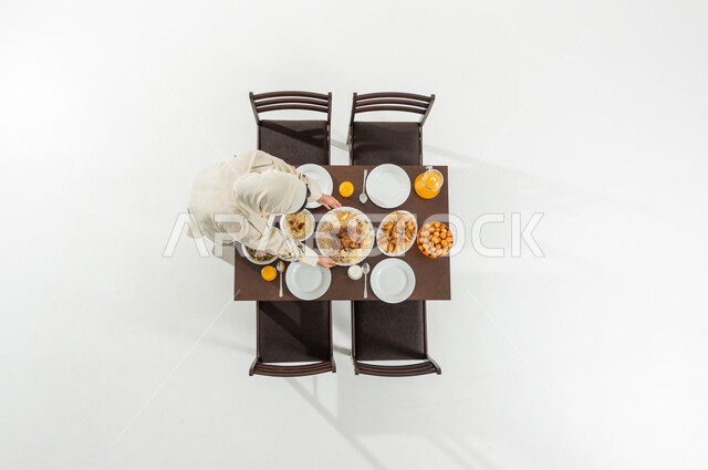 تجهيز سفرة الإفطار في شهر رمضان المبارك , بورتريه علوي عامودي لامرأة عربية خليجية سعودية ترتدي الثوب التقليدي و الحجاب تقوم بتحضير مائدة الطعام , أطباق شرقية متنوعة , أجواء رمضانية سعيدة, خلفية بيضاء