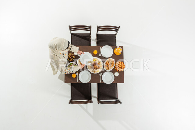 Iftar meal with various dishes, atmosphere of the holy month of Ramadan, vertical overhead portrait of a Saudi Gulf Arab woman wearing traditional dress and hijab preparing the dining table, a variety of foods, white background