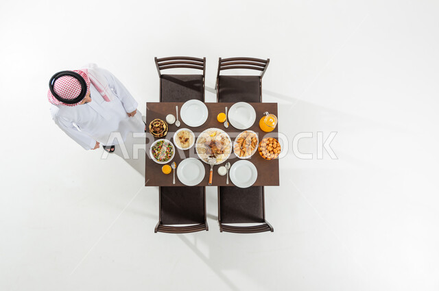 المساعدة في تحضير سفرة الإفطار , أجواء شهر رمضان المبارك , بورتريه علوي عامودي لرجل عربي خليجي سعودي يرتدي الثوب التقليدي و الشماغ يقف بجانب مائدة الطعام , مناسبات دينية إسلامية , خلفية بيضاء