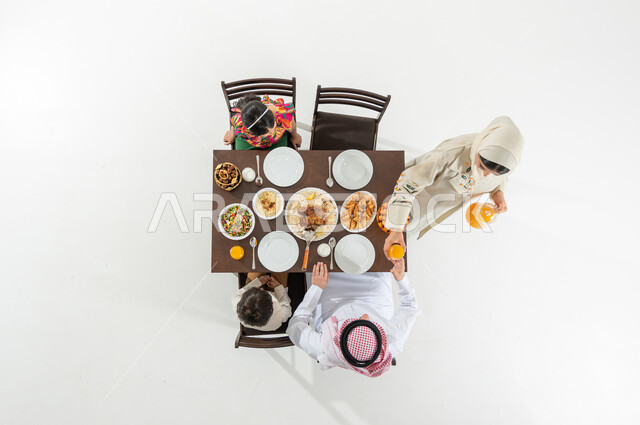 تقديم المشروبات الباردة على الإفطار , أجواء عائلية في شهر رمضان المبارك, بوتريه علوي عامودي لعائلة عربية خليجية سعودية يرتدون الثوب التقليدي يجلسون على مائدة الطعام , تشكيلة متنوعة من المأكولات , خلفية بيضاء