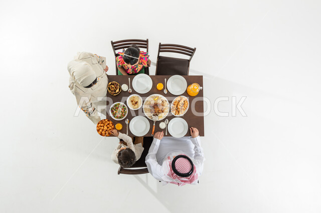 الاستمتاع بأكل الحلويات الشرقية , أجواء رمضانية عائلية , بوتريه علوي عامودي لعائلة عربية خليجية سعودية يرتدون الثوب التقليدي يجلسون على مائدة الطعام , سفرة مليئة بالأصناف المتنوعة , خلفية بيضاء