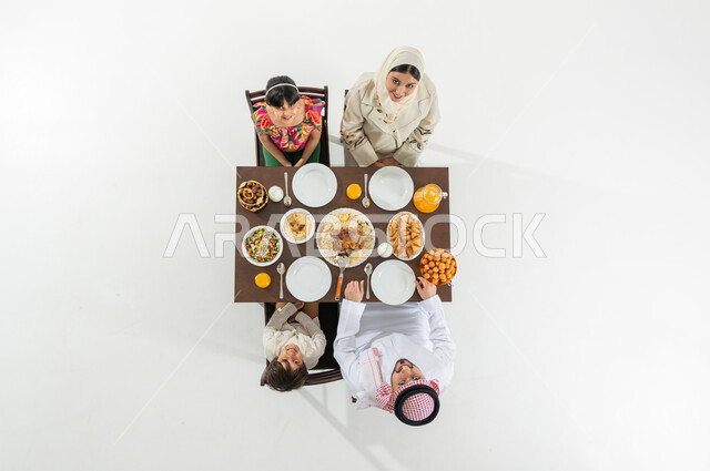 أجواء شهر رمضان المبارك , النظر إلى الكاميرا بإيماءات الفرح و السرور , بوتريه علوي عامودي لعائلة عربية خليجية سعودية يرتدون الثوب التقليدي يجلسون على مائدة الطعام يقومون بتناول الإفطار, تشكيلة متنوعة من المأكولات, خلفية بيضاء