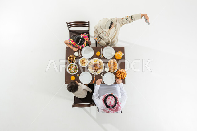 اجتماع العائلة في وقت الإفطار , أجواء شهر رمضان المبارك , بوتريه علوي عامودي لعائلة عربية خليجية سعودية يرتدون الثوب التقليدي يجلسون على مائدة الطعام , وجبات و أطباق شرقية, خلفية بيضاء