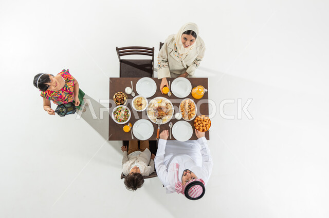 إفطار رمضاني عائلي , النظر إلى الكاميرا بإيماءات الفرح و السرور بشهر رمضان المبارك, بوتريه علوي عامودي لعائلة عربية خليجية سعودية يرتدون الثوب التقليدي يجلسون على مائدة الطعام , وجبات و أطباق شرقية , خلفية بيضاء