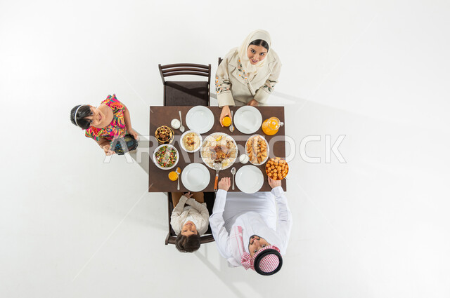 النظر إلى الكاميرا بإيماءات الفرح و السرور بشهر رمضان المبارك , بورتريه علوي عامودي لعائلة عربية خليجية سعودية يرتدون الثوب التقليدي يجلسون على مائدة الطعام يقومون بتناول الإفطار , تشكيلة متنوعة من المأكولات الشرقية , خلفية بيضاء