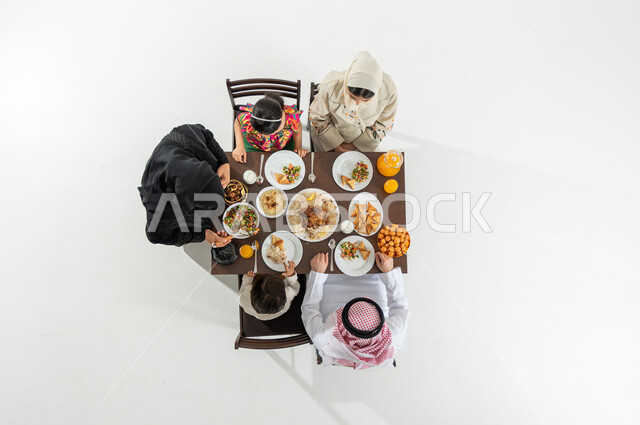 Serving oriental cuisine in Saudi Arabia, a vertical overhead portrait of a Saudi Gulf Arab family wearing traditional dress, sitting at the dining table eating breakfast, happy Ramadan atmosphere, white background