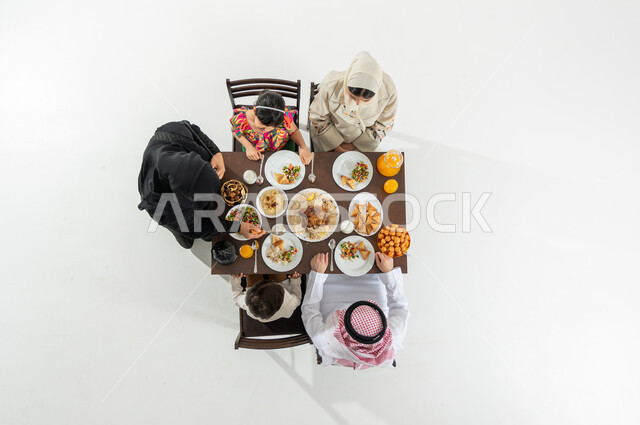 Enjoying spending iftar time with the family, serving oriental food, a vertical overhead portrait of a Saudi Gulf Arab family wearing the traditional dress sitting at the dining table, the atmosphere of the holy month of Ramadan, a white background