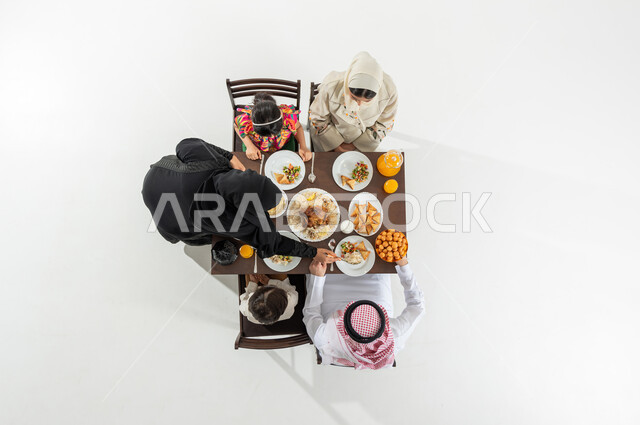تقديم المأكولات السعودية الشهية , أجواء رمضانية سعيدة , بورتريه علوي عامودي لعائلة عربية خليجية سعودية يرتدون الثوب التقليدي يجلسون على مائدة الطعام يقومون بتناول الإفطار , سفرة مليئة بالمأكولات المتنوعة , خلفية بيضاء
