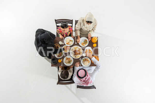 مفهوم الترابط و الأجواء الرمضانية , سكب الطعام لأفراد الأسرة , بورتريه علوي عامودي لعائلة عربية خليجية سعودية يرتدون الثوب التقليدي يجلسون على مائدة الطعام يقومون بتناول الإفطار , سفرة مليئة بالأصناف المتنوعة , خلفية بيضاء