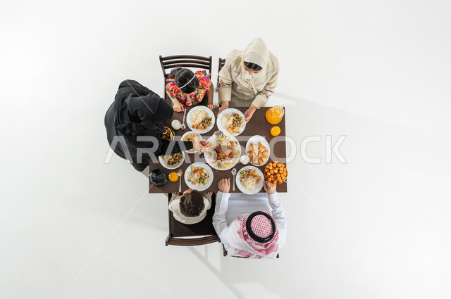 تقديم خدمات الطهي في السعودية , مشاركة الأسرة الإفطار , بورتريه علوي عامودي لعائلة عربية خليجية سعودية يرتدون الثوب التقليدي يجلسون على مائدة الطعام , تشكيلة متنوعة من المأكولات , خلفية بيضاء