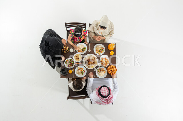 تقديم الأطعمة المتنوعة , أجواء رمضانية سعيدة , بورتريه علوي عامودي لعائلة عربية خليجية سعودية يرتدون الثوب التقليدي يجلسون على مائدة الطعام يقومون بتناول الإفطار , قضاء الأوقات الممتعة في شهر رمضان, خلفية بيضاء