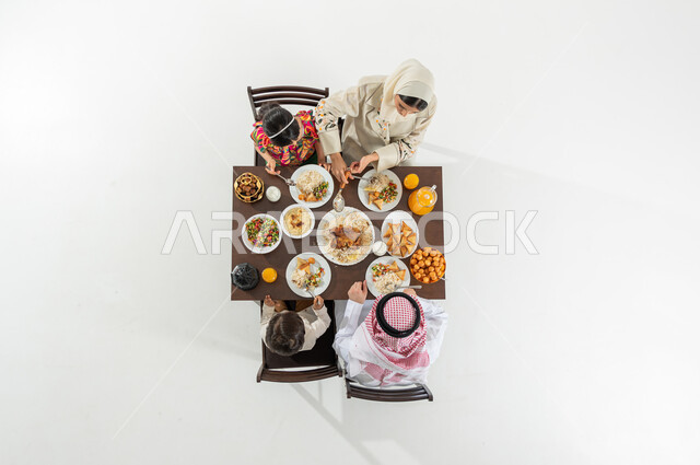 مفهوم المشاركة بين أفراد الأسرة , سفرة مليئة بالأصناف المتنوعة , بورتريه علوي عامودي لعائلة عربية خليجية سعودية يرتدون الثوب التقليدي يجلسون على مائدة الطعام يقومون بتناول الإفطار , قضاء الأوقات الممتعة مع الأسرة في شهر رمضان, خلفية بيضاء