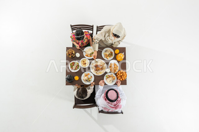 سفرة مليئة بالأصناف المتنوعة , مشاركة الإفطار مع الأسرة , بورتريه علوي عامودي لعائلة عربية خليجية سعودية يرتدون الثوب التقليدي يجلسون على مائدة الطعام يقومون بتناول الإفطار , قضاء أوقات ممتعة في شهر رمضان , خلفية بيضاء