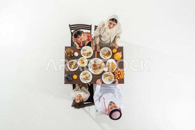 مشاركة الإفطار مع الأسرة , بورتريه علوي عامودي لعائلة عربية خليجية سعودية يرتدون الثوب التقليدي يجلسون على مائدة الطعام يقومون بتناول الإفطار , قضاء الأوقات الممتعة مع العائلة , النظر إلى الكاميرا بإيماءات الفرح و السرور , خلفية بيضاء