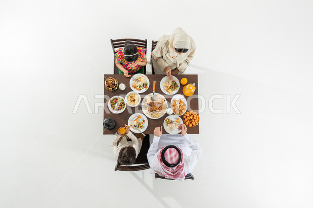 قضاء وقت الإفطار مع العائلة , أصناف متنوعة من المأكولات , بورتريه علوي عامودي لعائلة عربية خليجية سعودية يرتدون الثوب التقليدي يجلسون على مائدة الطعام يقومون بتناول الإفطار , أجواء رمضانية سعيدة , خلفية بيضاء