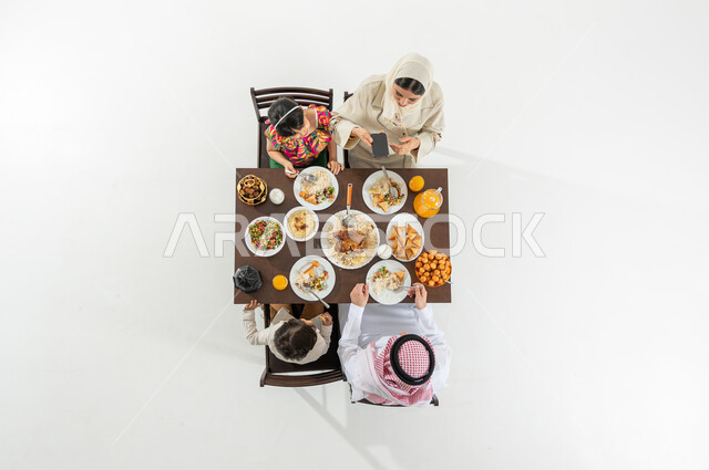 استخدام الهاتف المحمول لتصوير السفرة , أجواء رمضانية سعيدة , بورتريه علوي عامودي لعائلة عربية خليجية سعودية يرتدون الثوب التقليدي يجلسون على مائدة الطعام يقومون بتناول الإفطار , أصناف متنوعة من المأكولات ,خلفية بيضاء