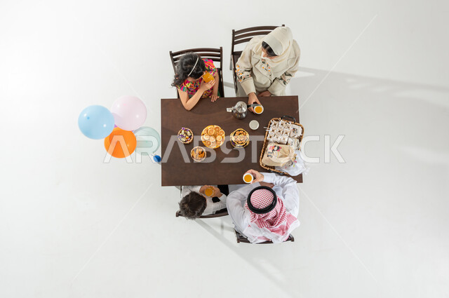 الاستمتاع بأجواء العيد مع الأسرة , بورتريه علوي عامودي لعائلة عربية خليجية سعودية يرتدون الثوب التقليدي يجلسون على مائدة الطعام يقومون بشرب العصير الطازج , مشروبات صيفية باردة , الاحتفال بالأعياد و المناسبات , خلفية بيضاء