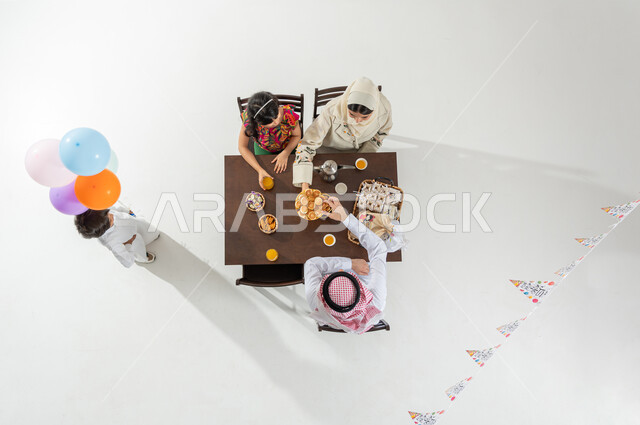 مشاركة حلويات العيد مع الأسرة , قضاء أوقات ممتعة مع العائلة , بورتريه علوي عامودي لعائلة عربية خليجية سعودية يرتدون الثوب التقليدي يجلسون على مائدة الطعام , أجواء الأعياد في السعودية, خلفية بيضاء