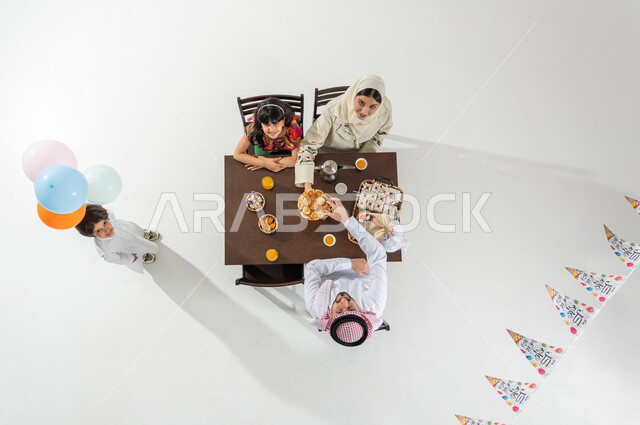 قضاء أوقات ممتعة مع الأسرة , بورتريه علوي عامودي لعائلة عربية خليجية سعودية يرتدون الثوب التقليدي يجلسون على مائدة الطعام , حلويات و مخبوزات العيد , النظر إلى الكاميرا بإيماءات الفرح و السرور , خلفية بيضاء