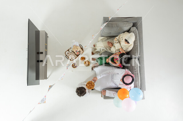 البهجة و السرور بقدوم العيد ,  بورتريه علوي عامودي لعائلة عربية خليجية سعودية يرتدون الثوب التقليدي يجلسون على الأريكة في غرفة المعيشة , أعياد و مناسبات إسلامية, تقديم الحلويات و المأكولات الشهية, عرض شاشة سوداء فارغة عبر التلفاز , خلفية بيضاء