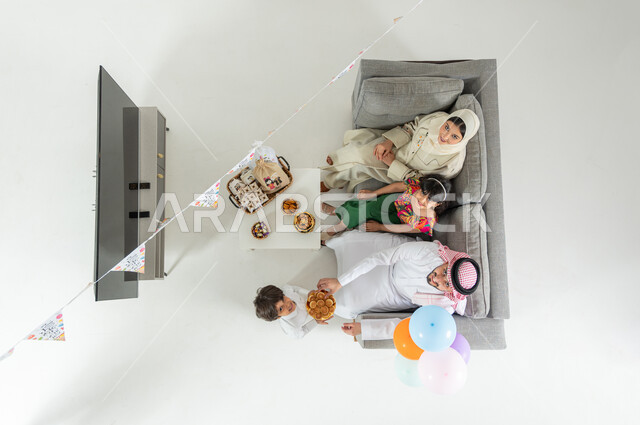 العادات و التقاليد في الأعياد و المناسبات , الاحتفال بقدوم العيد ,  بورتريه علوي عامودي لعائلة عربية خليجية سعودية يرتدون الثوب التقليدي يجلسون على الأريكة في غرفة المعيشة , مشاركة العائلة الحلوى و المأكولات الشهية, عرض شاشة سوداء فارغة عبر التلفاز , خلفية بيضاء