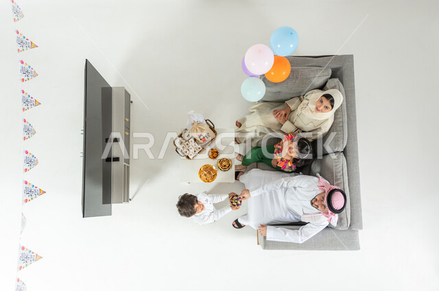 مشاركة العائلة الأجواء السعيدة , الاحتفال بقدوم العيد المبارك , بورتريه علوي عامودي لعائلة عربية خليجية سعودية يرتدون الثوب التقليدي يجلسون على الأريكة في غرفة المعيشة , إيماءات الفرح و السعادة , عرض شاشة سوداء فارغة عبر التلفاز , خلفية بيضاء