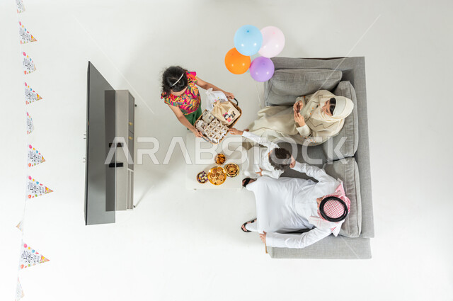 الاحتفال بقدوم العيد المبارك , بورتريه علوي عامودي لعائلة عربية خليجية سعودية يرتدون الثوب التقليدي يجلسون على الأريكة في غرفة المعيشة , تقديم الحلوى و المأكولات الشهية , أعياد و مناسبات إسلامية , عرض شاشة سوداء فارغة عبر التلفاز , خلفية بيضاء