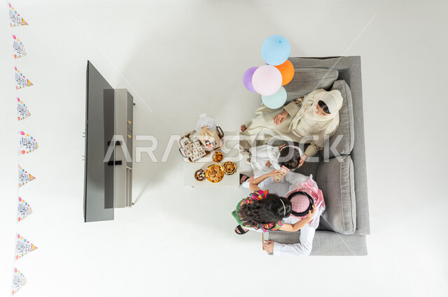 ارتداء ملابس العيد الجديدة , فتاة تقبل رأس والدها , بورتريه علوي عامودي لعائلة عربية خليجية سعودية يرتدون الثوب التقليدي يجلسون على الأريكة في غرفة المعيشة , مشاركة العائلة أجواء العيد , عرض شاشة سوداء فارغة عبر التلفاز , خلفية بيضاء